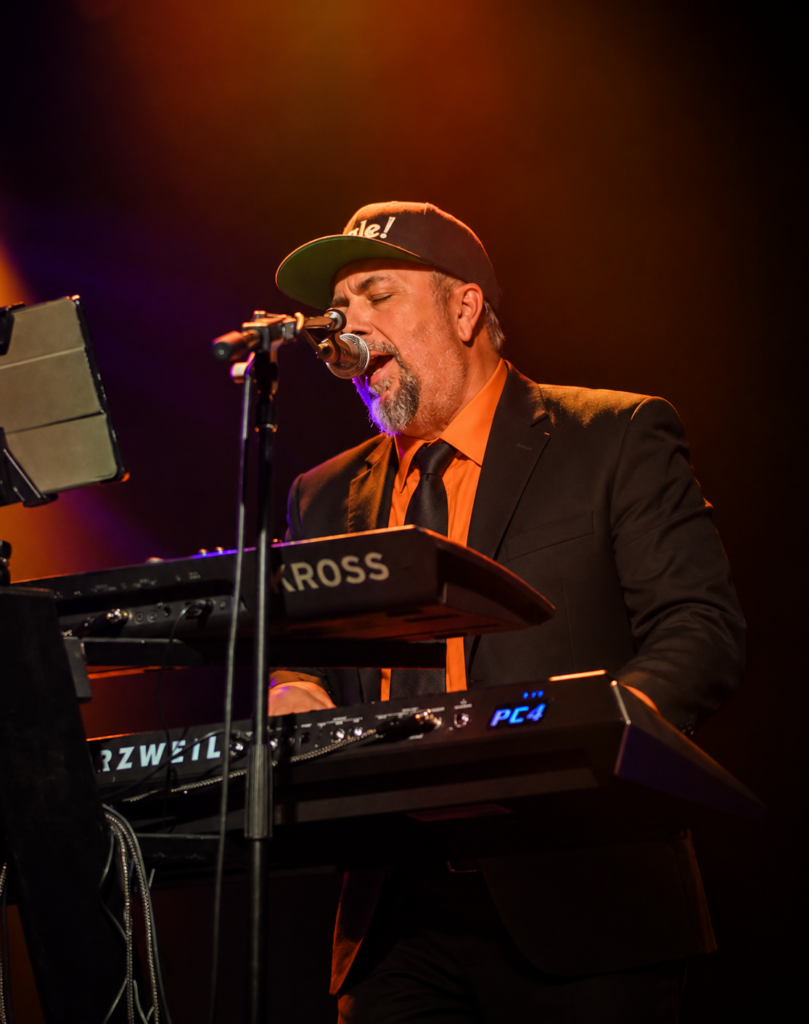 Órale Band keyboard player in an orange shirt and dark jacket at the keys